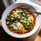 Spring peas and broad beans in a spicy tomato shakshuka, finished with crumbled feta, fresh herbs, and lemon wedges for brightness.