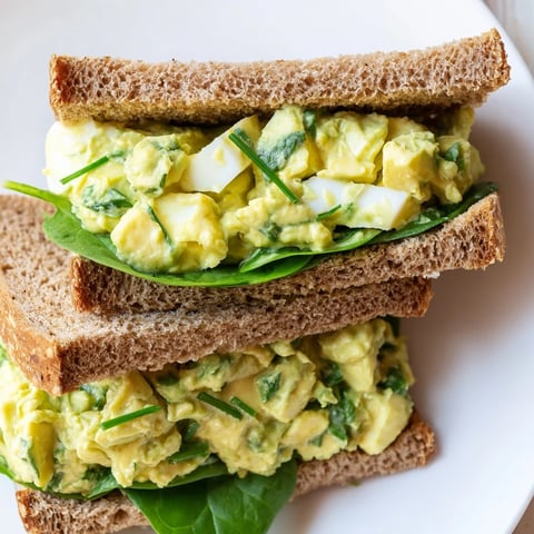 Golden toasted bread slices hold a hearty scoop of creamy avocado egg salad, with fresh spinach and tomato layered beneath.
