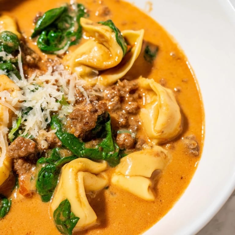 Close-up of Tuscan Tortellini Soup featuring cheese tortellini, crumbled sausage, and wilted spinach swimming in a rich, creamy broth.